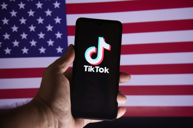 Mão segurado celular com logo do TikTok na tela,. em frente a uma bandeira americana