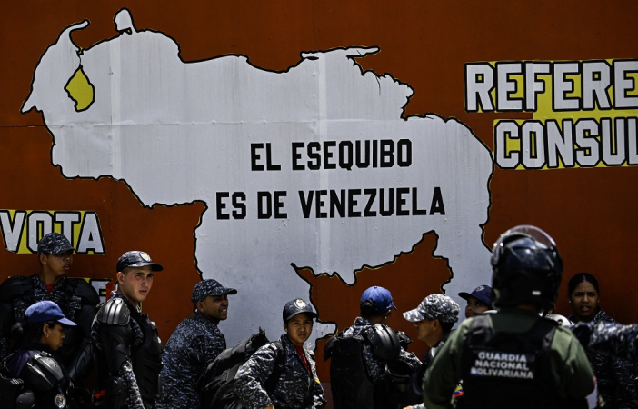 Integrantes da Polícia Nacional Bolivariana e da Guarda Nacional estão ao lado de um mural com o mapa da Venezuela e a frase "O Essequibo pertence à Venezuela" durante um protesto do Dia do Trabalhador em Caracas
