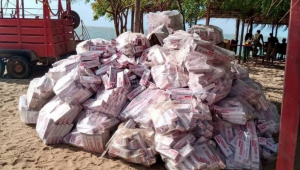 A imagem mostra uma grande pilha de sacos empilhados em uma praia, com areia ao redor. Os sacos são de cor clara e estão amarrados com fitas vermelhas. Ao fundo, há uma estrutura de palha que parece ser um quiosque, e algumas árvores verdes. Um veículo vermelho está visível à esquerda da imagem. Parte de carga de cigarros contrabandeados que estava em embarcação que encalhou em Cajueiro da Praia, litoral do Piauí
