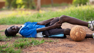 Jovem caído e com a mão no joelho em campo de futebol, ao lado da bola