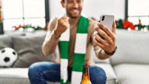Jovem hispânico apoiando partida de futebol usando smartphone sentado na decoração de natal em casa