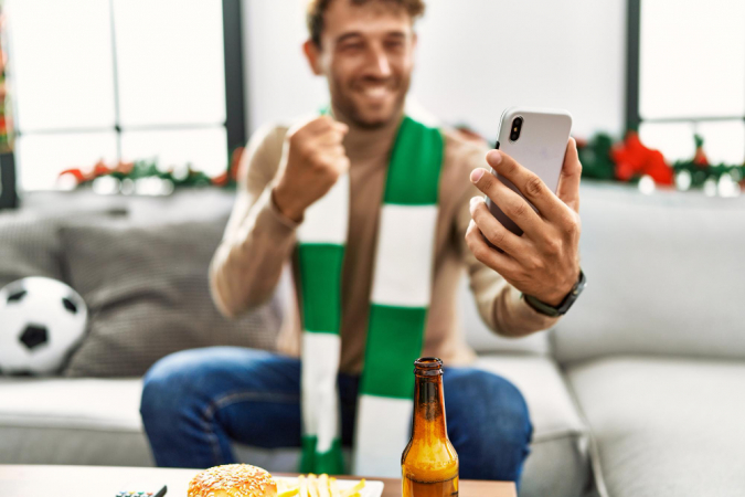 Jovem hispânico apoiando partida de futebol usando smartphone sentado na decoração de natal em casa
