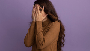 Linda garota assustada vestindo um suéter de gola alta marrom coberto com as mãos isoladas na parede roxa