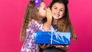 Menina satisfeita sussurra no ouvido de uma menina sorridente caucasiana segurando uma caixa de presente isolada na parede rosa