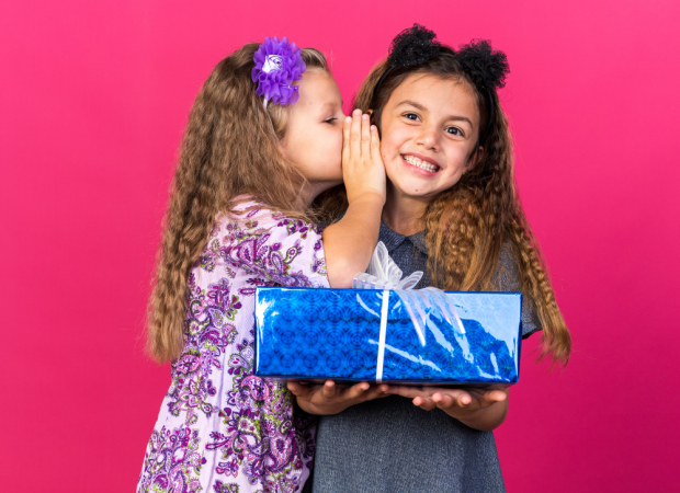 Menina satisfeita sussurra no ouvido de uma menina sorridente caucasiana segurando uma caixa de presente isolada na parede rosa