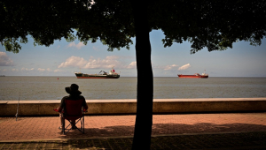 Um homem sentado em uma cadeira pesca enquanto o cargueiro Mistral (à esq.), que navega sob bandeira de Antígua e Barbuda, e o cargueiro Koole K, sob bandeira de São Vicente e Granadinas, permanecem ancorados na baía de Port of Spain