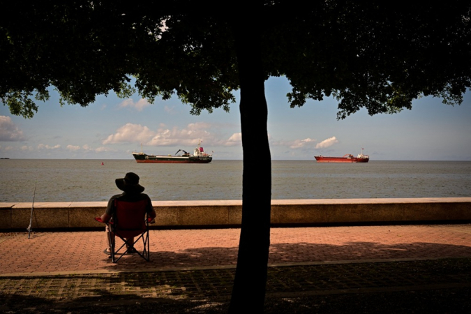 Um homem sentado em uma cadeira pesca enquanto o cargueiro Mistral (à esq.), que navega sob bandeira de Antígua e Barbuda, e o cargueiro Koole K, sob bandeira de São Vicente e Granadinas, permanecem ancorados na baía de Port of Spain