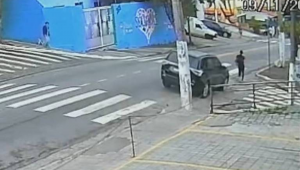 Homem está parado ao lado de carro preto estacionado próximo a esquina de rua com faixa de pedestres. Ambiente urbano com calçada e muro azul ao fundo. Momento em que mulher caminha pela calçada e veículo se aproxima segundos antes do atropelamento - Reprodução/TV globo