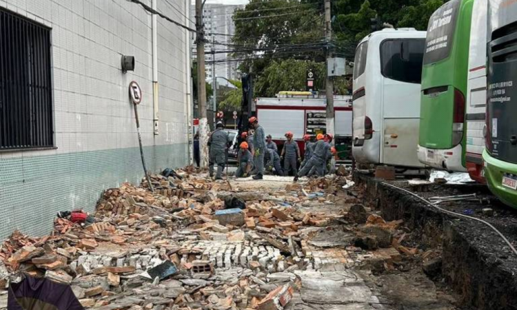Calçada destruída com concreto quebrado e entulho ao lado de ônibus verdes e brancos estacionados. Pessoas e caminhão ao fundo em dia nublado. Desabamento de muro no Brás deixa três vítimas - Corpo de Bombeiros