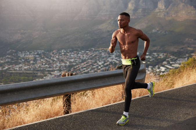 Homem de pele escura sem camisa corre rapidamente pela estrada, usa calçados esportivos e tem motivação para ser vencedor de competições de corrida
