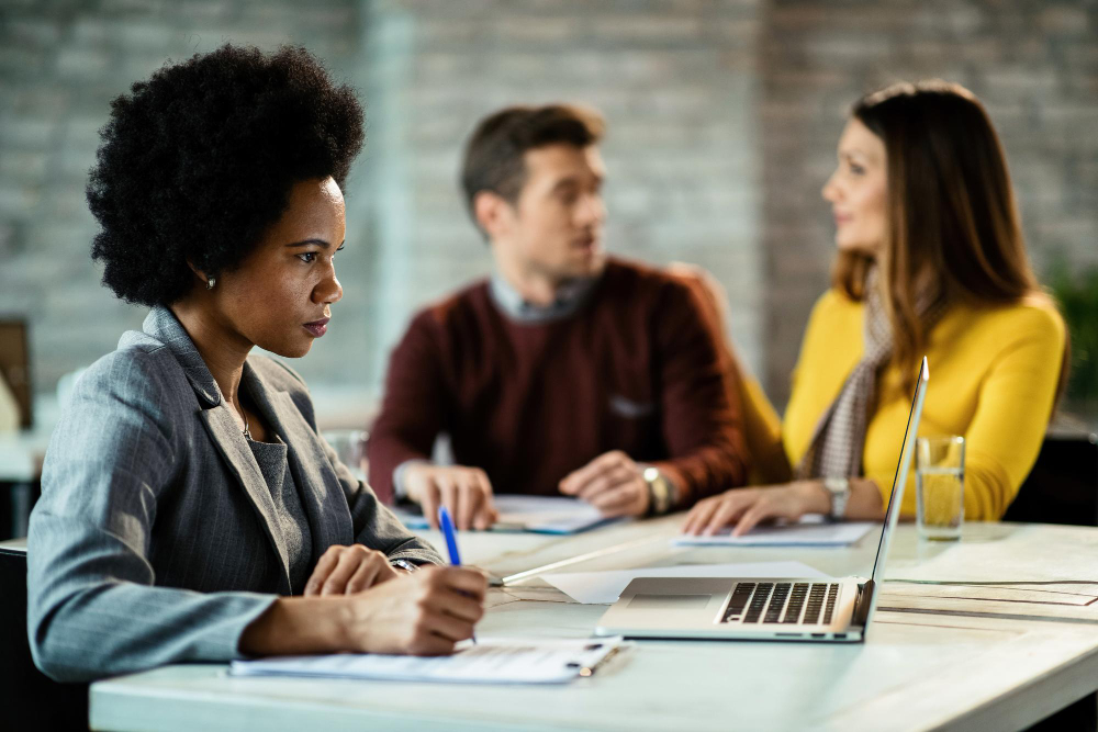Gerente de banco feminino negro usando laptop e escrevendo notas enquanto se encontra com um casal no escritório
