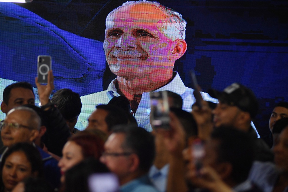 Uma tela de LCD exibe o retrato do candidato presidencial hondurenho Nasry Asfura, do Partido Nacional, enquanto apoiadores comemoram após o Conselho Nacional Eleitoral (CNE) declará-lo vencedor