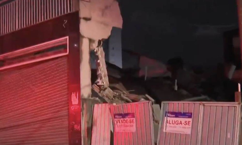 Prédio de quatro andares parcialmente desabado em área urbana de Guarulhos durante a noite. Fachada danificada com rachaduras visíveis e escombros no chão. Placas de 'Vende-se' e 'Aluga-se' fixadas em muro próximo. Iluminação vermelha de veículos de emergência ilumina a cena. Ambiente escuro com poucas construções ao redor. Prédio de quatro andares desaba em Guarulhos, na Grande São Paulo - Reprodução/Globonews