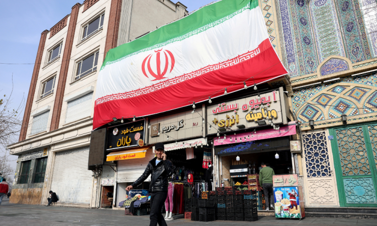 Um homem iraniano fala ao celular enquanto caminha ao lado de uma enorme bandeira nacional pendurada acima de lojas em Teerã, em 6 de fevereiro de 2026. O Irã e os Estados Unidos iniciaram negociações em Omã em 6 de fevereiro, com Washington se recusando a descartar uma ação militar contra a república islâmica por sua repressão mortal a protestos em massa.