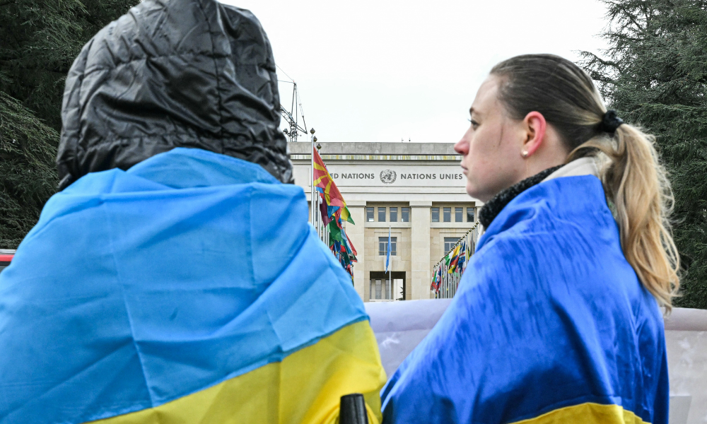 Manifestantes vestindo bandeiras ucranianas se posicionam em frente ao escritório das Nações Unidas enquanto uma nova rodada de negociações entre representantes russos, ucranianos e americanos, com o objetivo de encontrar uma solução para quatro anos de conflito na Ucrânia, ocorre em Genebra em 17 de fevereiro de 2026. (Foto de Harold CUNNINGHAM / AFP)