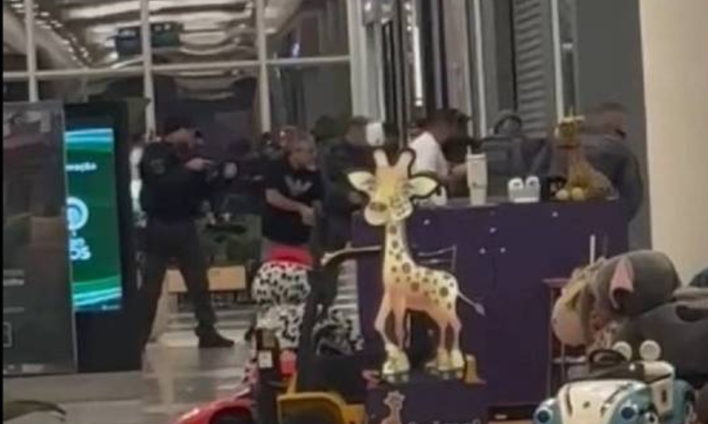 Grupo de pessoas em fila dentro de loja, com brinquedos expostos em prateleiras e no chão, incluindo um cavalo de balanço e carrinhos. Ambiente interno com iluminação artificial e estrutura metálica visível no teto.