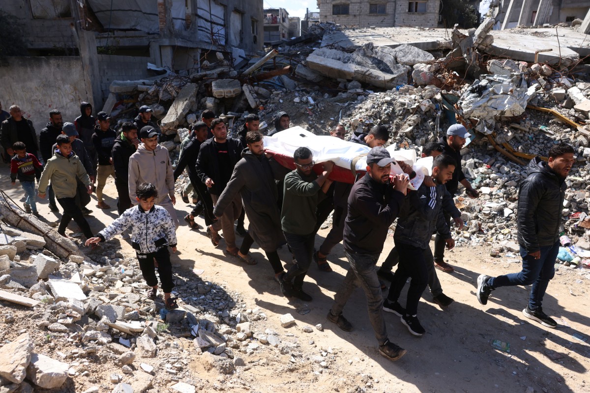 Palestinos carregam o corpo de uma pessoa morta em um ataque militar israelense, enquanto passam pelos escombros de um edifício destruído pelo exército de Israel, durante o funeral em Khan Yunis, no sul da Faixa de Gaza, em 27 de fevereiro de 2026