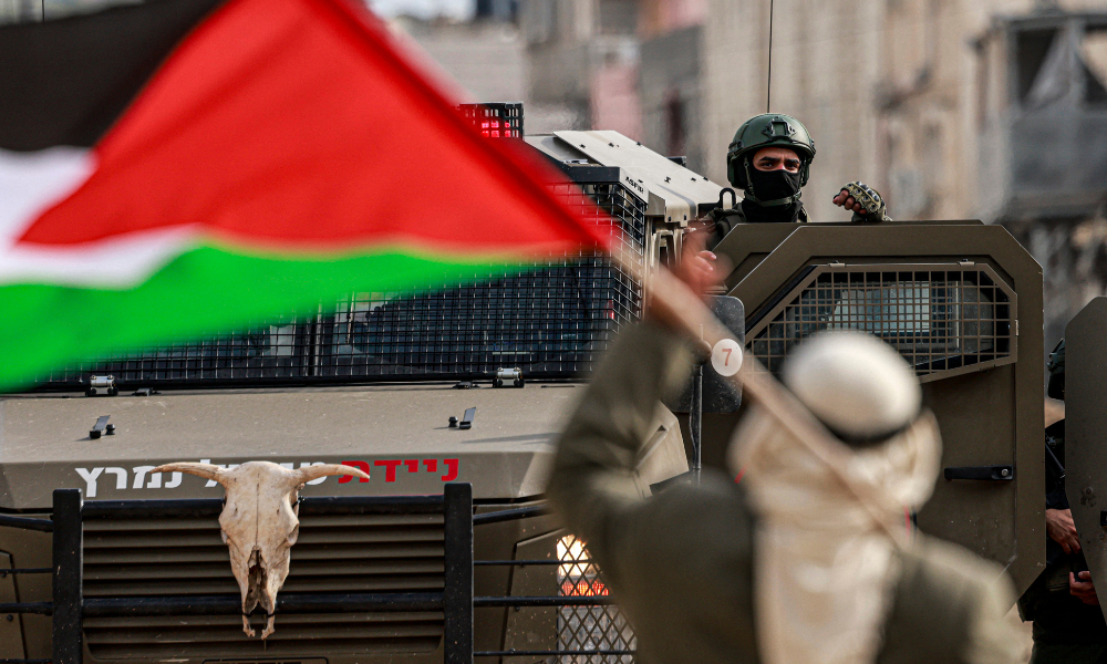 Um palestino segurando sua bandeira nacional gesticula em direção a um soldado israelense do lado de fora do campo de refugiados palestinos de Nur Shams, no norte da Cisjordânia ocupada por Israel, em 15 de dezembro de 2025, exigindo permissão para retornar à sua casa depois que o exército israelense o expulsou, juntamente com outros, no início de 2025, dando início a uma operação abrangente e ainda em andamento, que, segundo o governo israelense, visa erradicar grupos armados palestinos dos campos de refugiados no norte da Cisjordânia. O exército israelense demolirá 25 prédios residenciais, que abrigam até 100 casas familiares, no campo de refugiados de Nur Shams, no norte da Cisjordânia, ainda esta semana, informaram autoridades locais à AFP em 15 de dezembro de 2025. A operação militar nos campos de refugiados de Nur Shams, Tulkarem e Jenin deslocou todos os moradores e mais de 30.000 ainda não retornaram para casa.