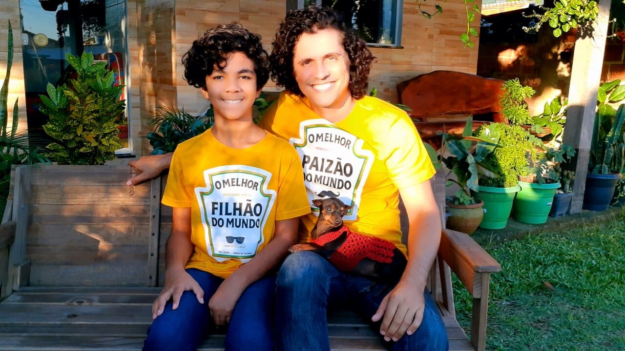 Pai e filho sentados em um banco com um cachorro pequeno no colo. Usam camisetas amarelas escritas 'Melhor filhão do mundo' e 'Melhor paizão do mundo'